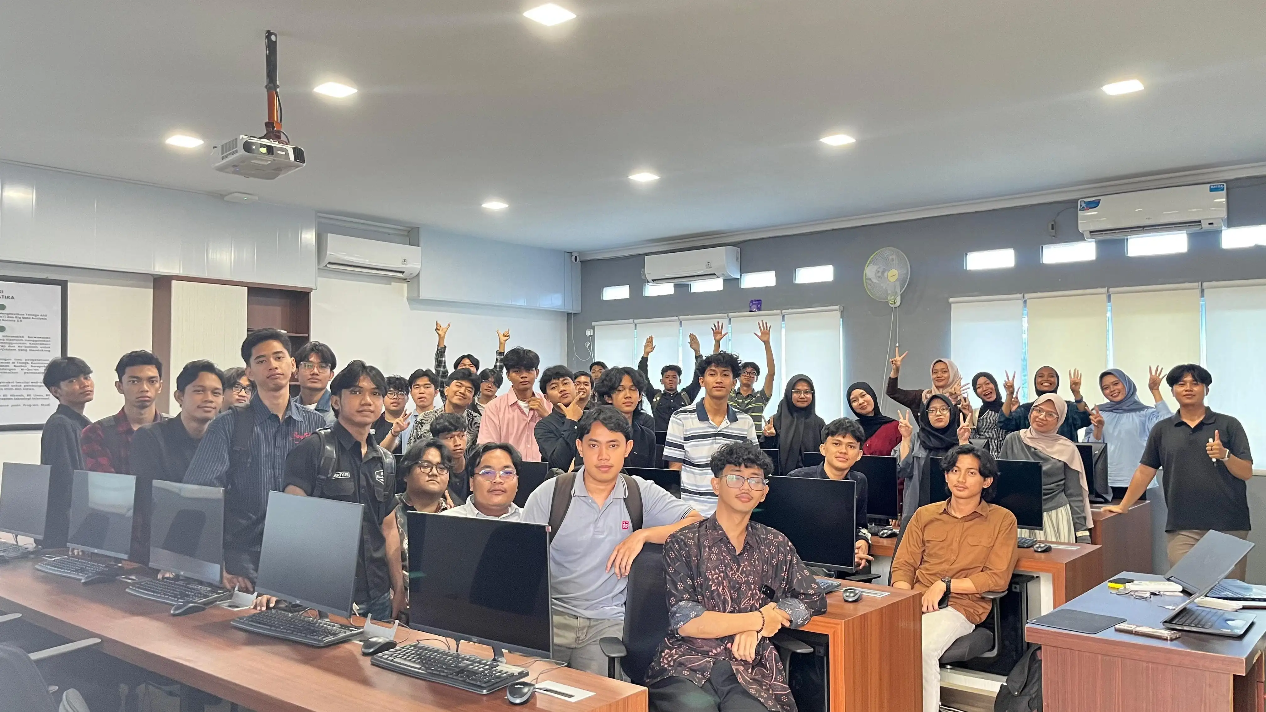 A computer lab at the end of a practicum session. Around
              thirty-five undergraduates stand and sit between two rows of
              monitors, facing the camera.